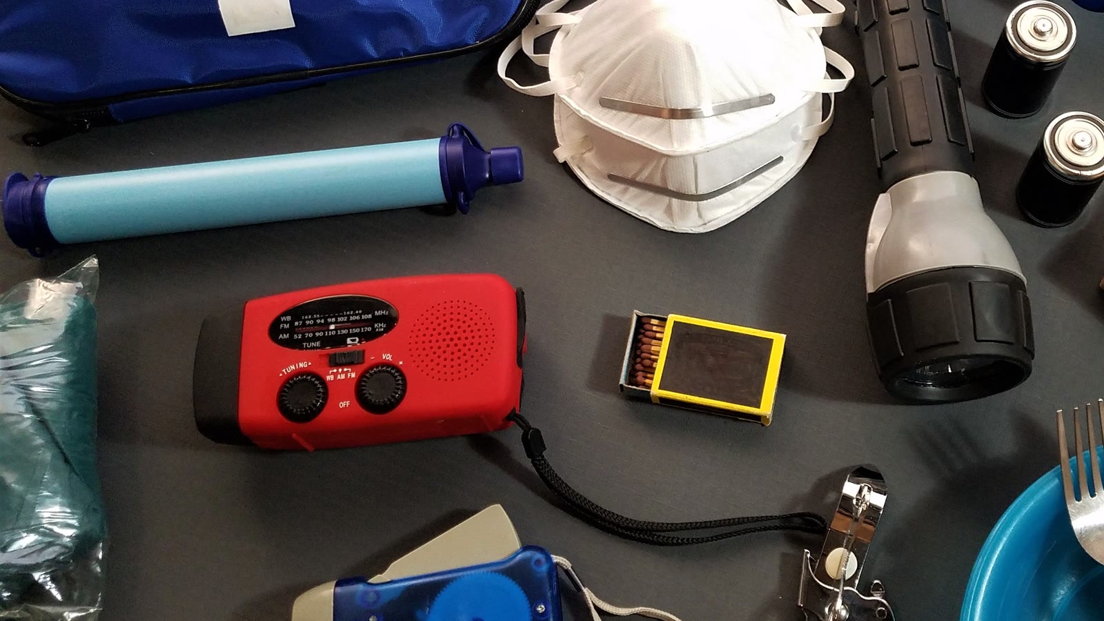 Håndsvingsradio, vandfilter, lommelygte, batterier, tændstikker og støvmasker lagt ud på et bord